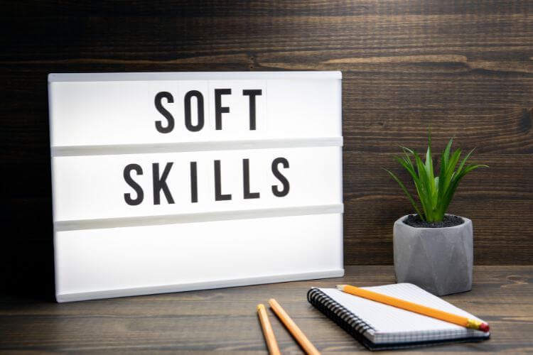 Lightbox sign with "Soft Skills" text, set on a wooden desk. Beside its are small potted plant, a notebook, and pencils.