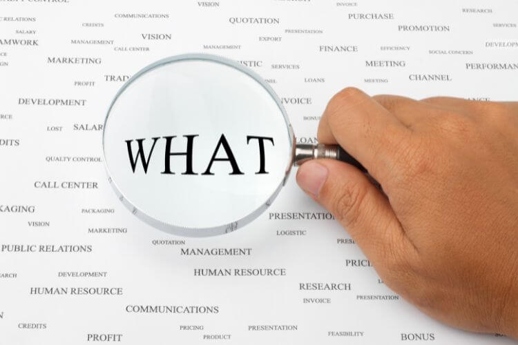 A hand holds a magnifying glass over a white sheet filled with business-related words. Write above letter "WHAT".